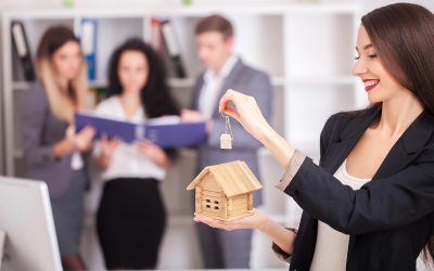 Real estate agent holding a miniature house and key, with clients discussing mortgage options in the background, emphasizing home ownership and financial guidance.