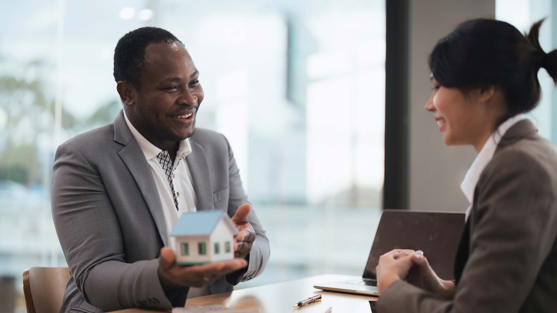 African American real estate agent presenting a miniature house model to a client in a modern office setting, illustrating personalized mortgage advice for Canadian homeowners.